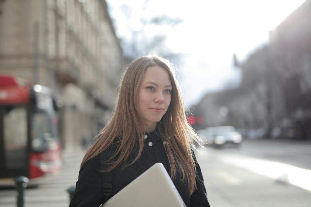 Confident young woman holding a laptop while walking on a bustling city street.